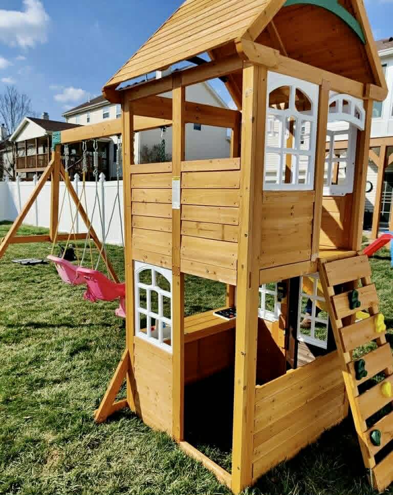 Two-level wooden playset with swings and climbing wall in backyard.